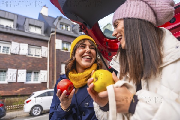 Two cheerful young women laughing while wearing hats and holding apples, standing by an open car trunk in an urban setting during cold weather, enjoying friendship