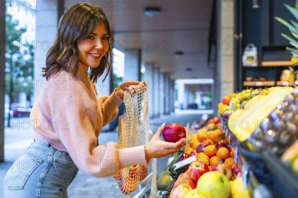 Young woman smiling while adding fresh red apples to her reusable mesh bag at an outdoor or street market, promoting healthy eating and sustainable consumer choices