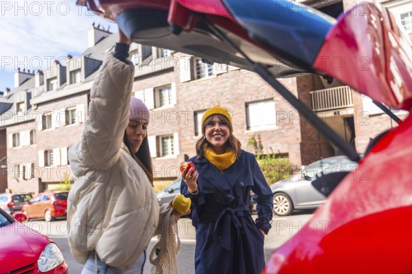 Two cheerful young women are holding apples and smiling, standing together by the open trunk of a red car in a city parking lot, enjoying a casual moment after shopping