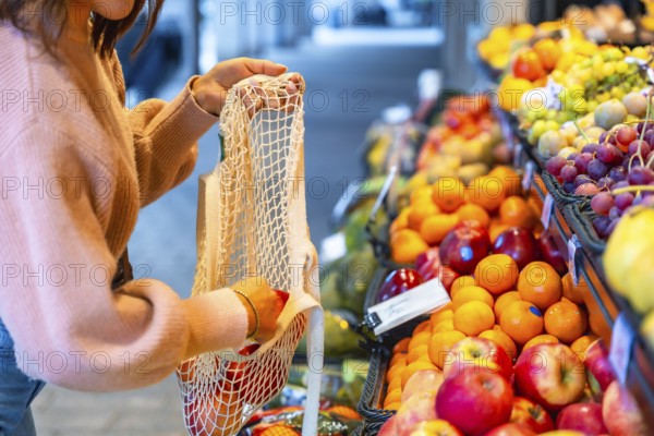 Woman selecting organic apples and oranges, placing them into a sustainable net shopping bag while grocery shopping at a vibrant fruit market, promoting eco friendly choices