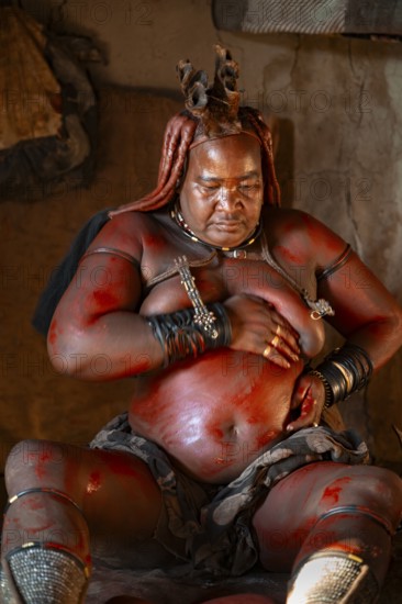 Elderly Himba woman rubbing herself with red paste for skin care, in a hut, traditional Himba village, Kaokoveld, Kunene, Namibia