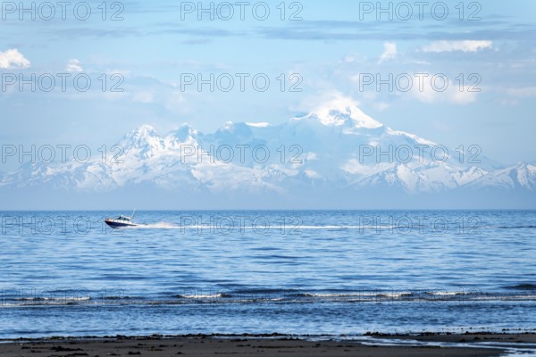 View over Cook Inlet to white mountain peaks of Mount Iliamna, motor boat rides on the ocean, mountains of the Aleutian Range, Anchor Point, Anchor River State Recreation Area, Alaska, USA