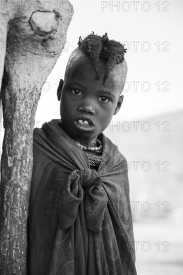 Black and white, Himba girl leaning at a traditional clay hut, in the morning, traditional Himba village, Kaokoveld, Kunene, Namibia