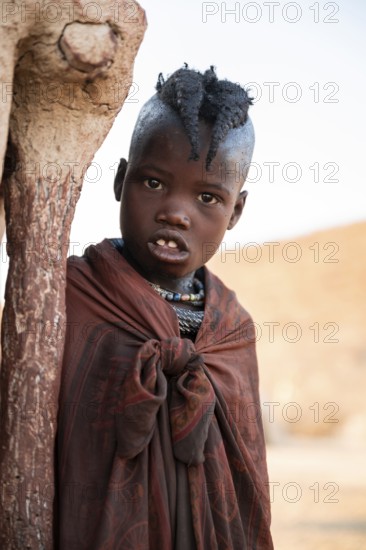 Himba girl leaning at a traditional clay hut, in the morning, traditional Himba village, Kaokoveld, Kunene, Namibia