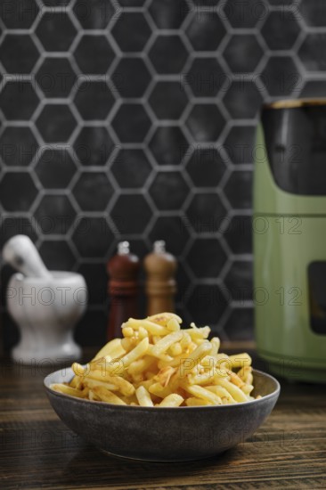 A bowl of crispy golden potato fries prepared in an air fryer is on a wooden kitchen table. Behind it are sleek salt and pepper shakers, and a modern kitchen appliance adds charm to the stylish decor