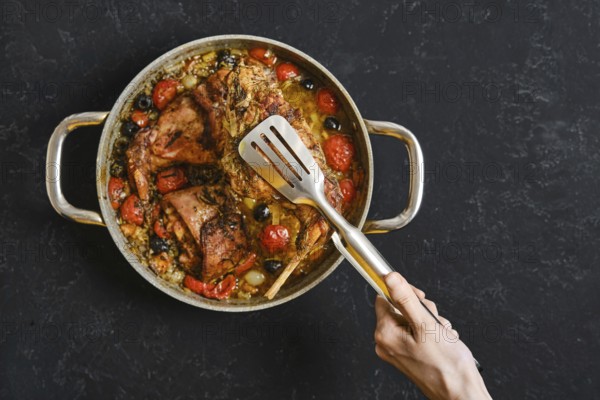 A hand uses a kitchen tongs to lift a tender rabbit leg from a pan filled with roasted tomatoes and olives. This dish is a delightful centerpiece for a cozy meal, rich in flavors and colors