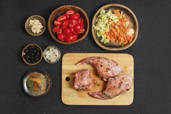 A rabbit's hind legs and a saddle are placed on a wooden cutting board, surrounded by fresh ingredients like tomatoes, carrots, and herbs