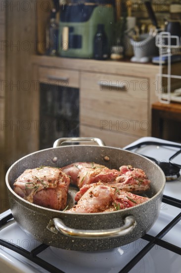 A rabbit's hind leg and saddle is simmering in a skillet, seasoned with herbs, on a gas stove in a warm kitchen. The rustic surroundings and wooden cabinets create a homey atmosphere