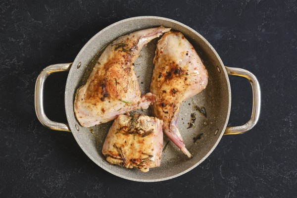 Two succulent rabbit hind legs and a saddle are cooked in a skillet. They are seasoned with herbs and spices, showcasing a golden-brown color