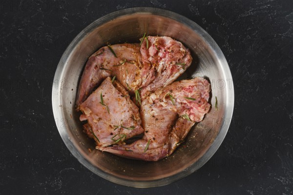 Two raw hind leg and saddle of rabbit is marinating with herbs and spices in a metal bowl. It is prepared for cooking. This scene captures the essence of preparing a meal
