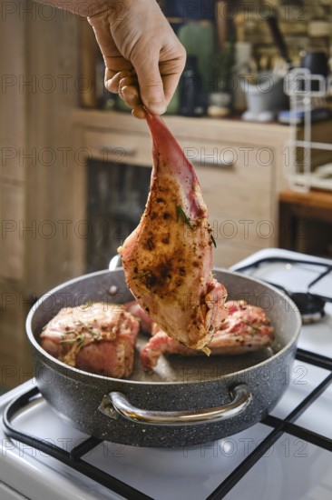 A person holds a marinated rabbit hind leg above a pot with other meat cooking inside. The kitchen has wooden cabinets and a warm atmosphere, indicating a cozy meal preparation time
