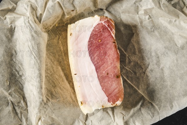 A slice of cured meat lies on a piece of parchment paper in a kitchen. The meat features a mix of pink meat and white lard, showcasing its quality and texture. Fresh herbs surround its edges