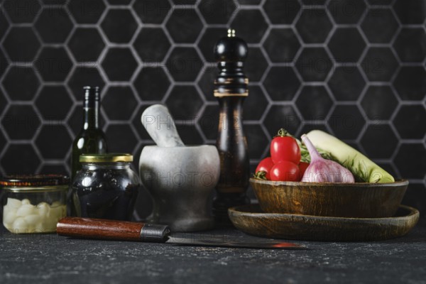 A variety of fresh vegetables such as tomatoes, onions, and greens sit on a dark kitchen counter alongside cooking tools