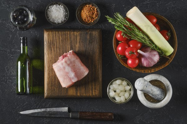 Fresh ingredients are beautifully arranged on a wooden board, including a piece of raw rabbit saddle meat, ripe tomatoes, garlic, leeks, olives, and herbs, ready for cooking a flavorful dish at home