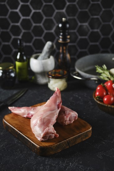 Raw rabbit hind legs on a wooden cutting board in a well-equipped kitchen. Fresh vegetables, oils, and various kitchen tools surround the meat, indicating preparation for a dish