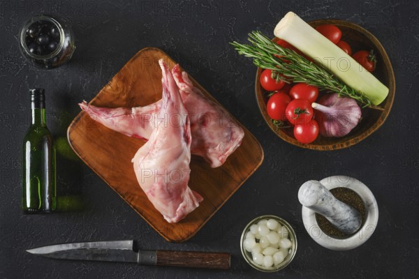 Top view of two raw rabbit hind legs are on a wooden board surrounded by fresh vegetables, herbs, and seasonings. The scene captures ingredients for a flavorful meal preparation on a dark surface