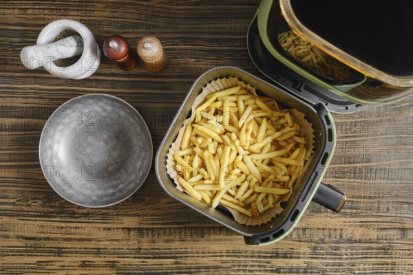 Golden fries are freshly cooked in an air fryer, resting in a basket lined with paper. A ceramic plate sits nearby, and salt and spice shakers are positioned on the wooden table