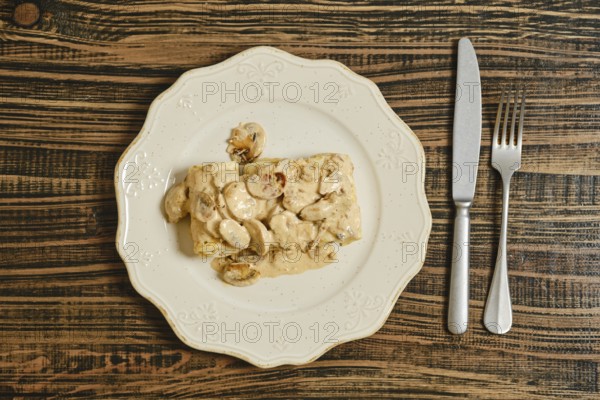 Crepes filled with seasoned meat, topped with a rich and creamy mushroom sauce. The dish is elegantly presented on a white plate against a wooden table