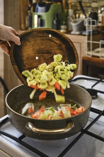 Sliced leeks and carrots are being poured from a wooden bowl into a pan on the stove. The warm light and homey setting create an inviting atmosphere for cooking a nutritious dish