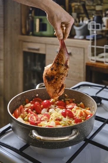A hand holds a seasoned rabbit leg above a pot filled with fresh vegetables, including tomatoes and onions, simmering on the stove. The warm kitchen provides an inviting cooking atmosphere