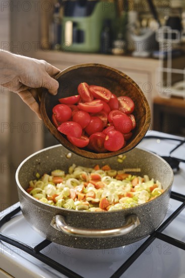 A person is adding chopped red tomatoes from a wooden bowl into a pot that is already filled with sauteed vegetables
