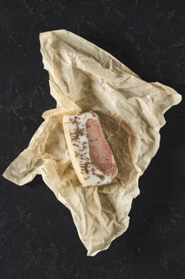 A rich piece of cured meat is neatly wrapped in crumpled brown paper resting on a dark surface. This traditional delicacy showcases a blend of flavors, ready for tasting or cooking
