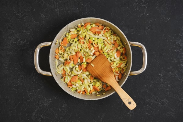 Chopped carrots and leeks are cooking in a large pan on a dark surface. A wooden spoon stirs the colorful vegetables