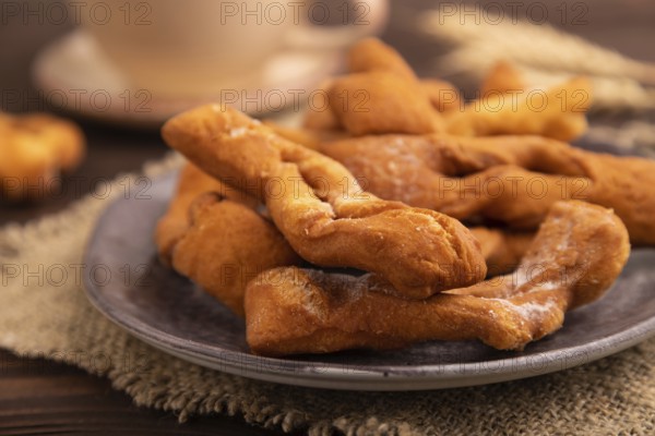 Crunchy biscuit Brushwood cookies sprinkled with powdered sugar on brown wooden background and linen textile, cup of coffee, side view, close up, selective focus