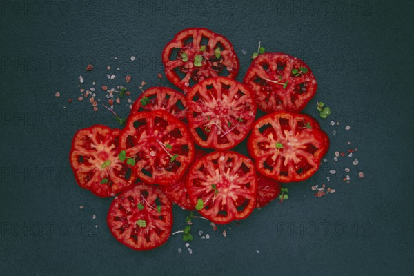 Freshly sliced ripe tomatoes arranged on a textured dark surface with herbs and salt scattered around