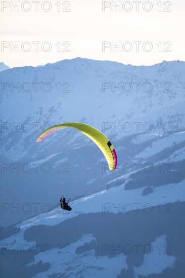 Paragliders flying over snowy mountain peaks in winter in evening light, Kitzbühel Alps, Tyrol, Austria