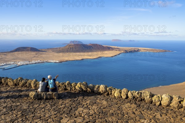 Tourists on a bench on the viewing platform at the Mirador del Río viewpoint, in the evening light, view of the sea and of the island of La Graciosa with volcanic craters, designed by artist César Manrique, Lanzarote, Canary Islands, Spain