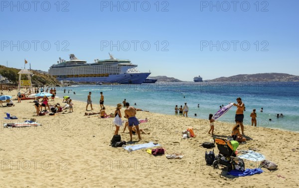 Mykonos, Cyclades, Greece - Voyager of the Seas cruise ship is located in Mykonos Harbour Bay. In front, tourists and locals bathe on the sandy beach of Paralia Agios Stefanos