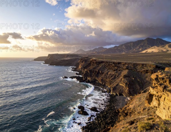 Desert landscape converges with ocean along a rugged coastal area, shore break at cliffs, blue cloudy sky at sunset, dry landscape in an aerial drone view, AI generated
