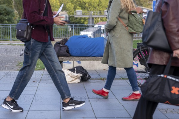 Man sleeping on a bench, travelling bag serves as pillow, in the city centre, passers-by walk by