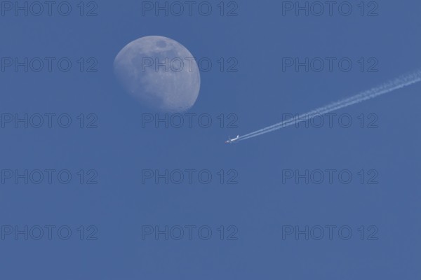 Boeing 737 jet passenger aircraft of Norweign Air with a vapour trial or contrail behind flying in a blue sky passing by the moon, England, United Kingdom