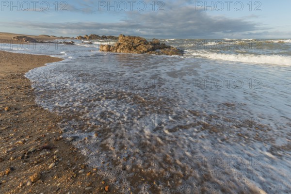 A wide sandy beach stretches out with scattered pebbles and beach shells. Gentle waves crash against the shore under a partially cloudy sky during late afternoon. Plage de l'Aubraie, Les Sables d'Olonne, Vendee, France