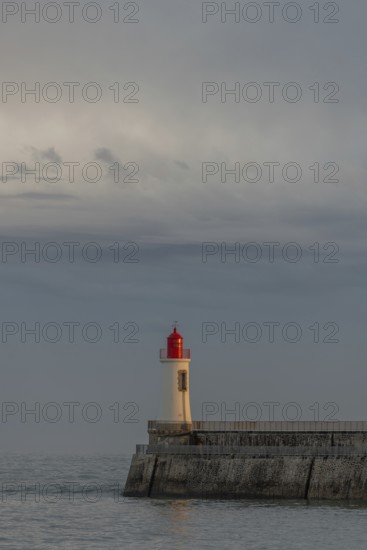 A lighthouse stands on a pier and casts its light over calm waters at dusk. The sky is filled with dramatic clouds, adding to the tranquil atmosphere of the coastal landscape. Les Sables d'Olonne, Vendee, France