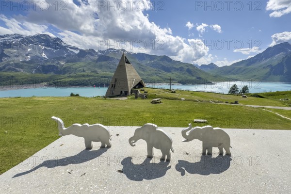 View from viewing platform to Mont-Cenis Pyramid Museum with stylised pyramid Museum in memory of Egypt campaigns of Emperor Napoleon Bonaparte and time in front of construction of reservoir, behind it Lac du Mont Cenis, in front on parapet small stylised figures of elephants Symbols for crossing of Alps Crossing of Alps by Hannibal, Mont Cenis Massif, Massif du Mont-Cenis, Graian Alps, Lanslebourg, Rhone-Alpes, France