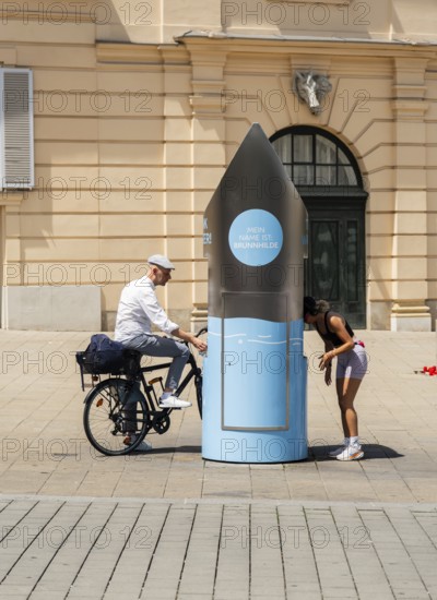 Two people in front of a drinking water fountain in the city centre of Vienna, Austria