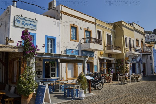 Row of historic buildings with restaurants and flowers on cobblestone street in sunshine, Agia Marina, Leros, Dodecanese, Greek Islands, Greece