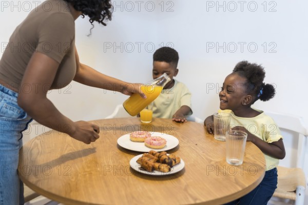 Mother pouring orange juice for her two children during breakfast, with donuts and pastries on the table