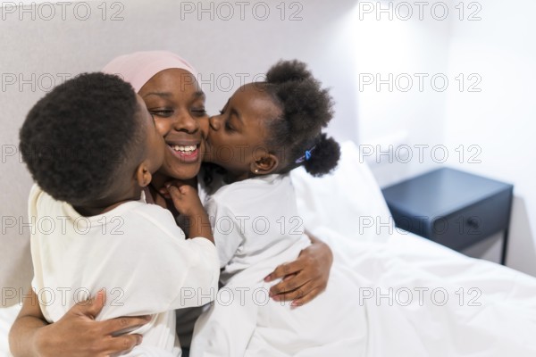 Two children kissing their smiling mother with cancer in bed, enjoying family time together