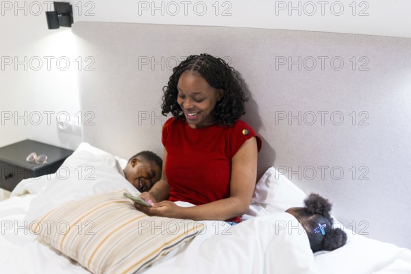 Smiling mother using smartphone while her two children are sleeping in bed, enjoying a quiet moment in the bedroom