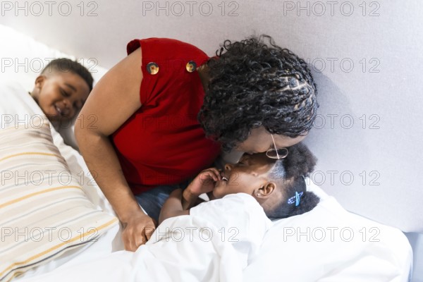 Mother kisses her daughter goodnight while putting her to bed, as her brother watches from the other side of the bed