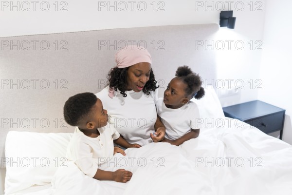 Mother undergoing cancer treatment, wearing a headscarf, sharing a heartwarming moment with her two children in bed, highlighting family support during challenging times