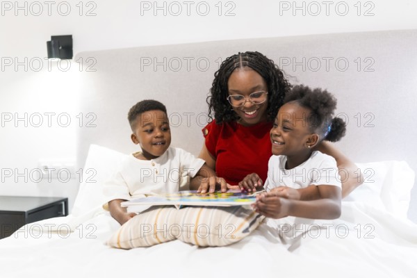 Smiling mother sharing a bedtime story with her two joyful children while cozily nestled on the bed, fostering love and connection