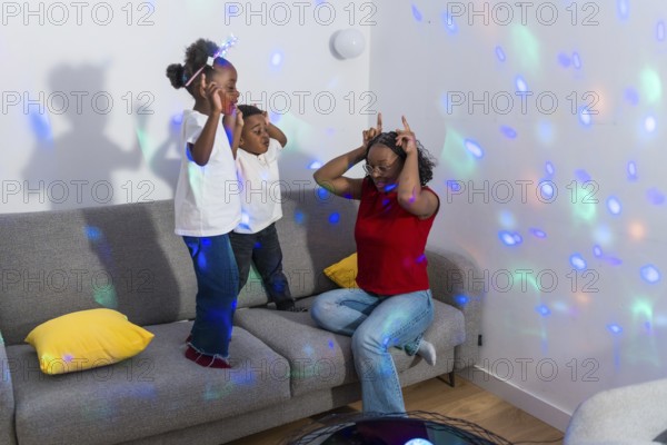 Mother making funny faces while kids dancing joyfully on the sofa, illuminated by colorful disco lights in a lively living room