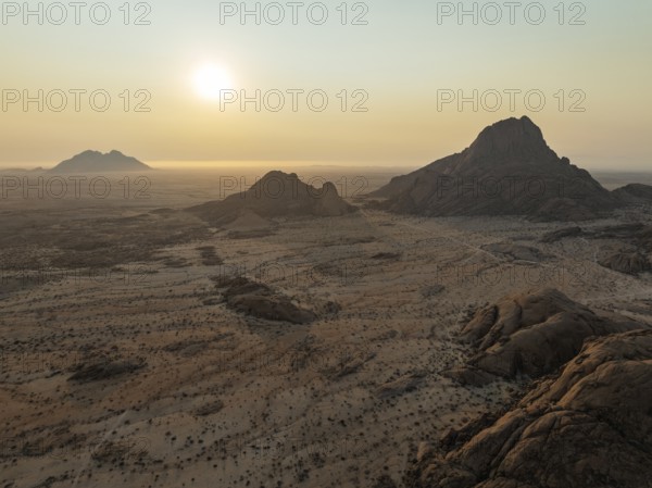 The isolated Spitzkoppe mountain (1728 m) - on the right - majestically rises above the surrounding desert plains. In the distance the coast and the Atlantic Ocean. At sunset. Aerial view. Drone shot. Damaraland, Namibia