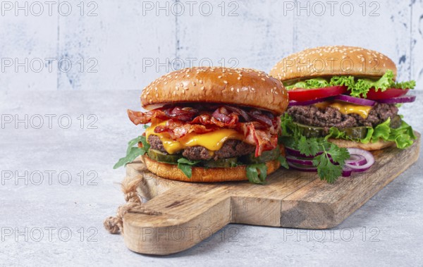 Two burgers with beef patty, fried bacon, cheddar cheese, tomatoes, on a light table