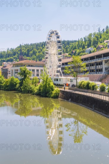 Ferris wheel reflected in the calm river, surrounded by buildings and wooded hills, Calw, Germany